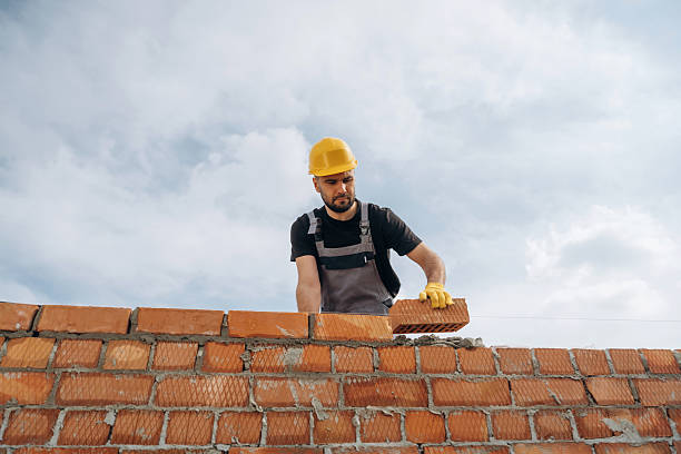 Against cloudy sky, making wall. Construction worker is at job with bricks outdoors.