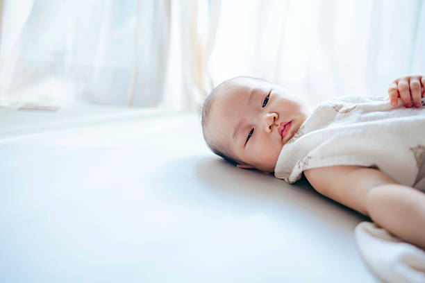 An Asian baby girl lies comfortably on a soft bed,  The scene captures the importance of early sensory development and reflects the attentive care modern parents provide to meet their baby’s health, safety, and emotional needs. It also subtly highlights how today’s families, under economic pressure and an abundance of information, seek trusted products and resources to support mindful parenting.