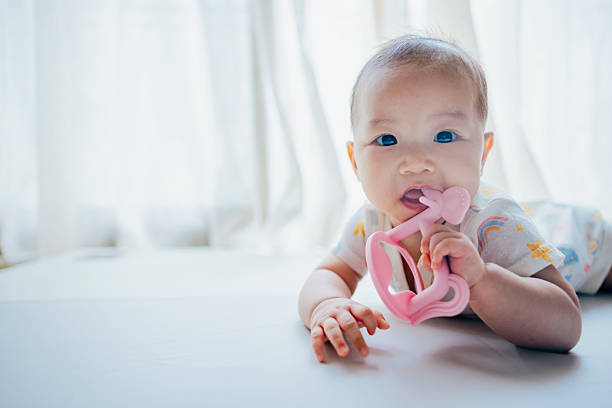 An Asian baby girl lies comfortably on a soft bed, playing with a teether while showing a focused and joyful expression. The scene captures the importance of early sensory development and reflects the attentive care modern parents provide to meet their baby’s health, safety, and emotional needs. It also subtly highlights how today’s families, under economic pressure and an abundance of information, seek trusted products and resources to support mindful parenting.