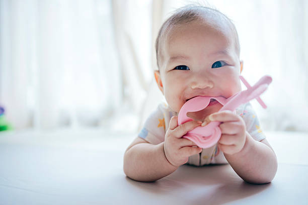 An Asian baby girl lies comfortably on a soft bed, playing with a teether while showing a focused and joyful expression. The scene captures the importance of early sensory development and reflects the attentive care modern parents provide to meet their baby’s health, safety, and emotional needs. It also subtly highlights how today’s families, under economic pressure and an abundance of information, seek trusted products and resources to support mindful parenting.
