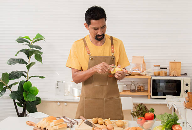 istock Asian man with beard sitting makes sandwich and divide two peice with knife in kitchen for breakfast in morning. Male wear apron enjoy make food for health care in house, healthy food at home. 2211912708
