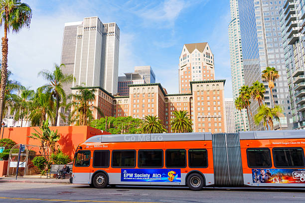 Los Angeles, California - December 3, 2024: Vibrant, orange metro bus stops at Pershing Square, surrounded by skyscrapers and palm trees, showcasing urban life in Los Angeles on a sunny day.
