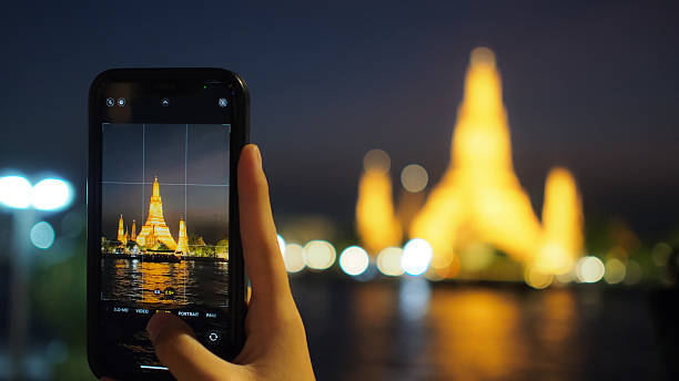A hand captures Wat Arun at night using a smartphone, with glowing golden lights reflecting on the water. Perfect for travel, technology, and culture-themed content.