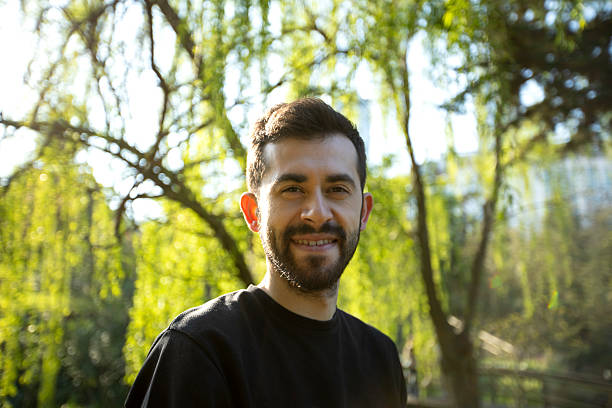 istock A smiling man with a beard stands outdoors under bright sunlight, surrounded by lush green trees. He is wearing a black shirt and looks happy and relaxed in the natural setting. 2211959479