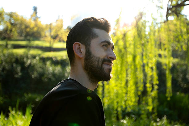 istock A smiling man with a beard stands outdoors under bright sunlight, surrounded by lush green trees. He is wearing a black shirt and looks happy and relaxed in the natural setting. 2211959710
