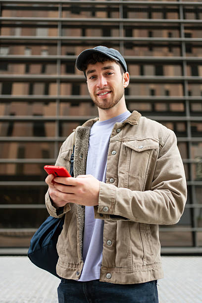 istock Smiling student with casual cap using mobile phone app in front of college campus building 2211967699