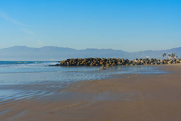 Rock jetty along Venice Beach with gentle waves washing ashore and distant mountains visible under a clear blue sky.