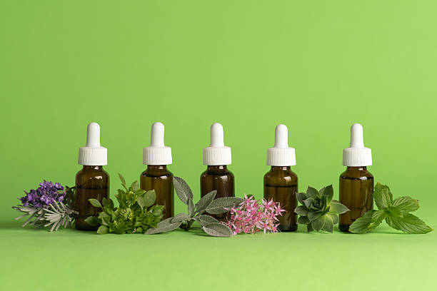 Five amber dropper bottles with white caps are elegantly arranged alongside a vibrant mix of fresh herbs and delicate flowers, set against a bold green backdrop. This image evokes a sense of natural beauty, wellness, and organic skincare, perfect for promoting eco-friendly health and beauty products.