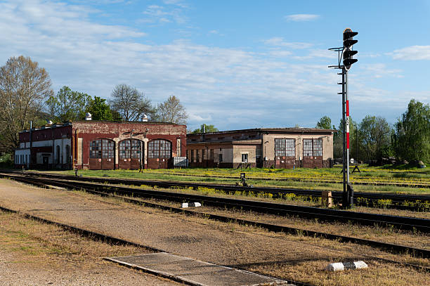 An old railway depot with weathered brick buildings and arched windows sits beside multiple train tracks. A tall signal post stands in the foreground under a partly cloudy sky, evoking a nostalgic railway atmosphere.