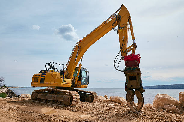 istock Yellow tracked excavator with hydraulic claw attachment working on a rocky coastal construction site near the sea under a partly cloudy sky 2211981899