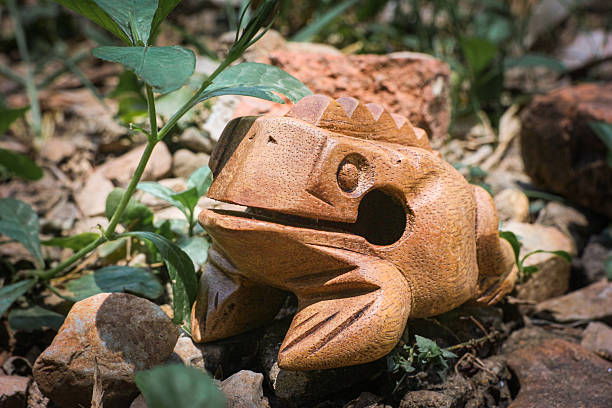 A large , three dimensional wooden frog sculpture hiding under lush green leaves, captured in a tropical rainforest simulation with soft sunlight filtering through the foliage.
