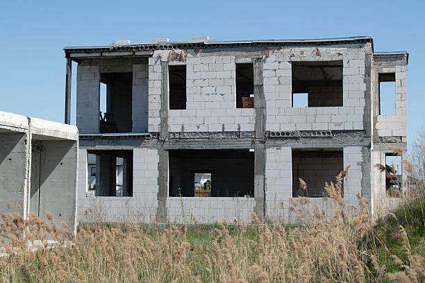 istock An unfinished building stands neglected at a construction site, with empty windows and cracked walls. Tall grass grows around the structure under a clear blue sky 2211989087