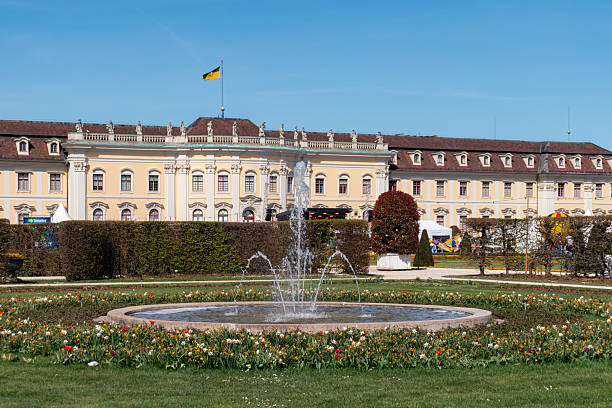 A decorative stone fountain sprays arcs of water in a lush green park on a sunny day. The light catches the droplets mid-air, creating a sparkling effect, while a vintage-style streetlamp and distant pedestrians add to the classical and serene atmosphere of the scene.