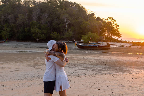 A couple shares a playful hug on a sandy beach at sunset, with a traditional wooden boat in the background. Relaxed tropical vibe.