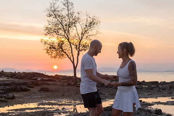 A couple holds hands and smiles at each other by the ocean during a colorful sunset, with a tree in the background.
