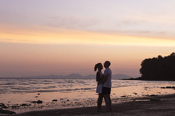 A romantic silhouette of a couple embracing on a tropical beach at sunset, with gentle waves in the background.