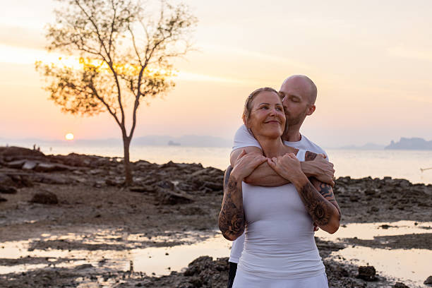A couple shares a loving embrace on rocky beach terrain during sunset, with a lone tree and ocean in the background.