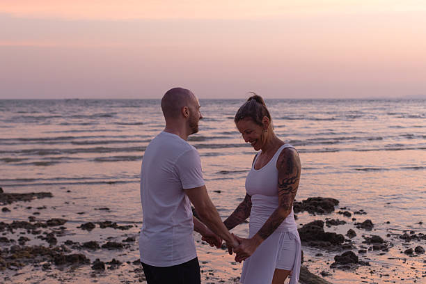 A couple stands close, holding hands tenderly on a rocky beach during a peaceful tropical sunset.