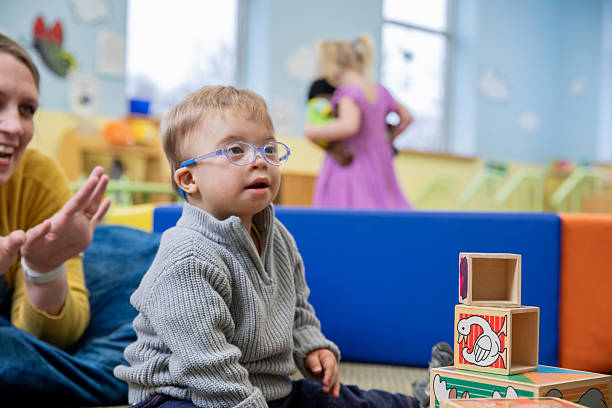 istock Toddler boy who has Down Syndrome is in classroom is stacking blocks with help from special education teacher 2212027265
