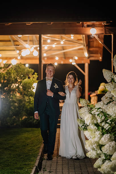 istock Bride and groom walk arm-in-arm at night under glowing lights, smiling joyfully, surrounded by lush white flowers and a romantic outdoor atmosphere. 2212037986