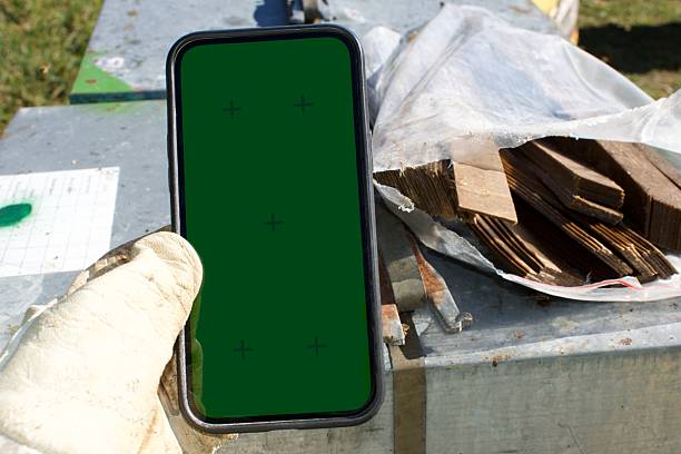 A mobile phone device held in the hand of a beekeeper in a rural beehive landscape in the background are beehives. The mobile phone technology is in focus while the beehives are  out of focus. This is a personal perspective concept for Beekeeping with Technology and Data Input. The mobile phone is in green screen.