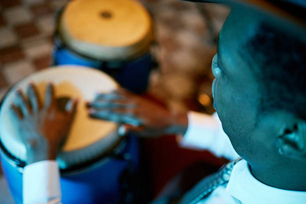 Over shoulder shot of Black man playing conga drums while performing Afro Cuban jazz along with music band in bar under vibrant neon lights