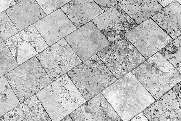 Rustic black and white image of square stone tiles with uneven texture laid diagonally outdoors, cobbled road