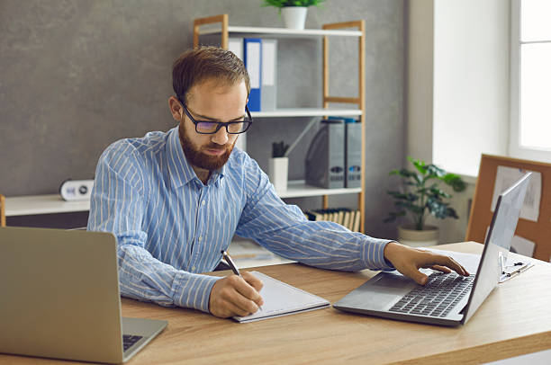 istock Serious concentrated man sitting at desk with laptops and taking notes in notebook 2212068312