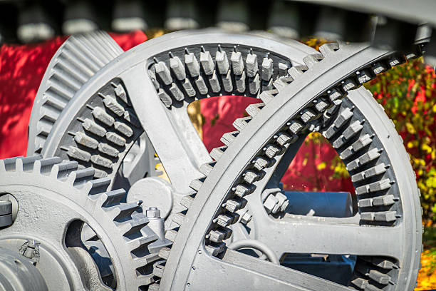 Close-up of large metallic gears engaged in a mechanical process, highlighting intricate design and functionality. Sunlight casts shadows, emphasizing the textures and precision of the machinery.