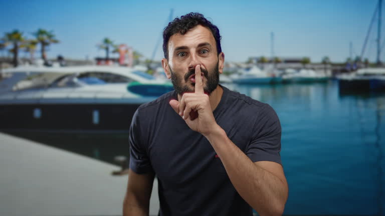 Hispanic man with a beard gestures for silence in a sunny port setting with yachts and palm trees in the background, emphasizing a serene maritime atmosphere.
