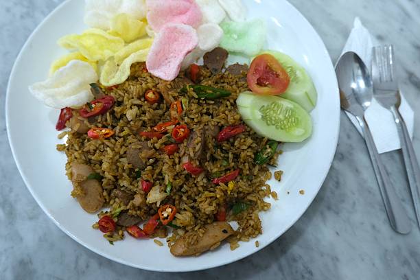 Top view of Indonesian fried rice or nasi goreng served with colorful crackers, cucumber, tomato slices, and cutlery on a marble table