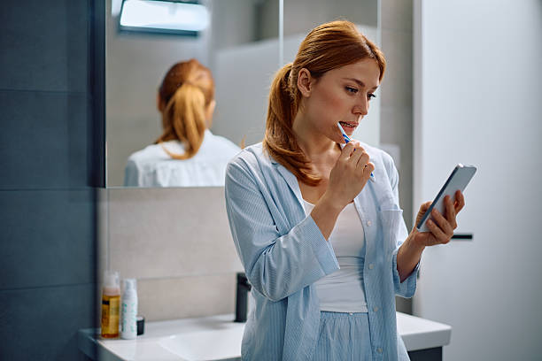 istock Mid adult woman reading text message on cell phone while brushing teeth in the bathroom. 2212083151