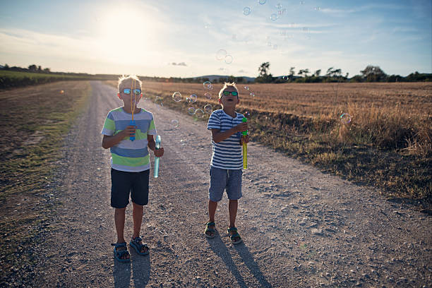 Two young boys enjoying a carefree afternoon blowing bubbles on a rural dirt road under a bright summer sky. Both boys wear sunglasses and casual striped shirts, surrounded by golden fields and the warm glow of the setting sun. The playful bubbles float through the air.Shot with Nikon D850