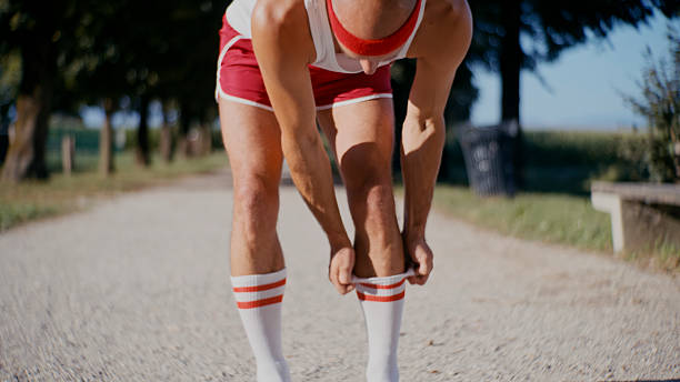 Close-up portrait of mid adult Caucasian male athlete bending forward to adjust white athletic socks with red stripes, wearing red running shorts on rural dirt path.