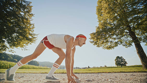 A mid-adult Caucasian man with a mustache wearing a retro outfit prepares to run on a sunny rural path surrounded by lush greenery and trees.