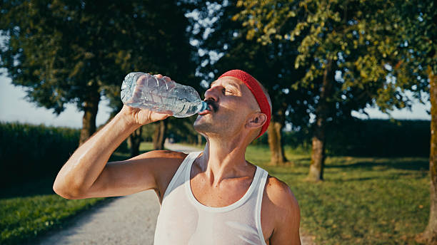 Close-up portrait of mid adult Caucasian man in red baseball cap and white tank top drinking from plastic water bottle on sunny park path surrounded by trees.