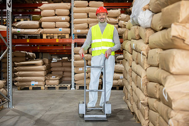A male warehouse worker stands with a dolly and smiles, reflecting positivity and professionalism in a busy industrial environment.