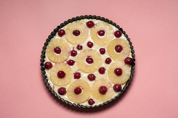 A vibrant unbaked sweet pie topped with pineapple slices and bright cherries, beautifully set against a soft pink background, showcasing a cheerful and colorful preparation scene before baking.