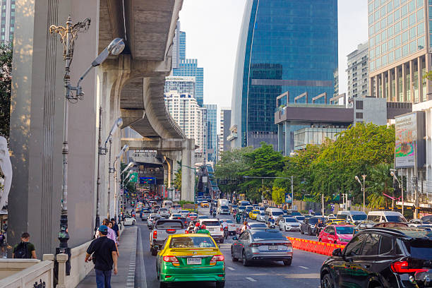 View along ruling traffic jam and rush hour on Phaya Thai Road in Bangkok downtown with view from south top north. Traffic jam is in both directions. A few people and pedestrians are on sidewalk at left side. BTS skytrain line and bridge is above street at left area. In right and cengter background are modern office building skyscrapers.