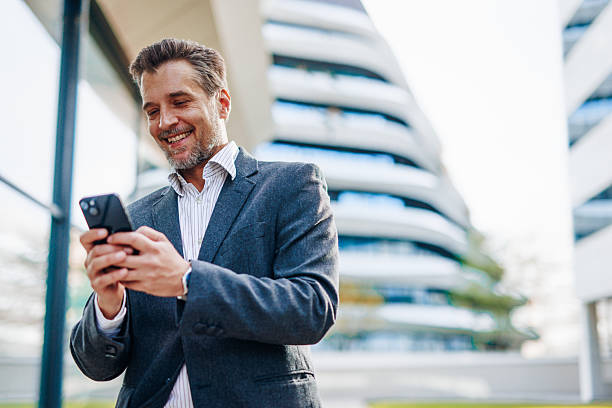 istock Smiling businessman using smartphone in front of modern office buildings 2212123073
