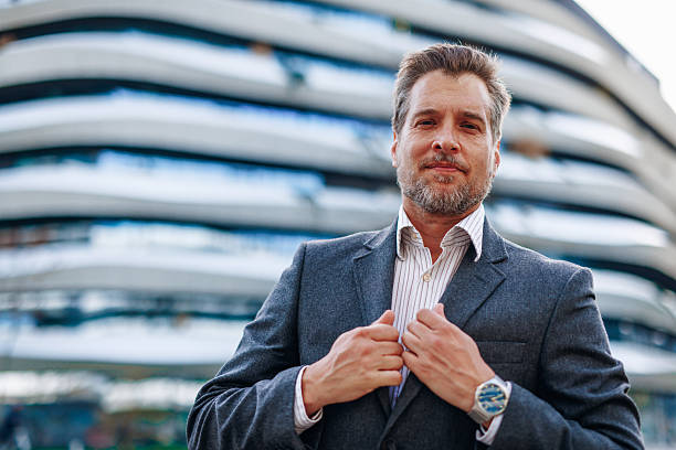 Portrait of a confident mature businessman adjusting his suit while standing in front of a modern office building, exuding professionalism and success in the corporate world
