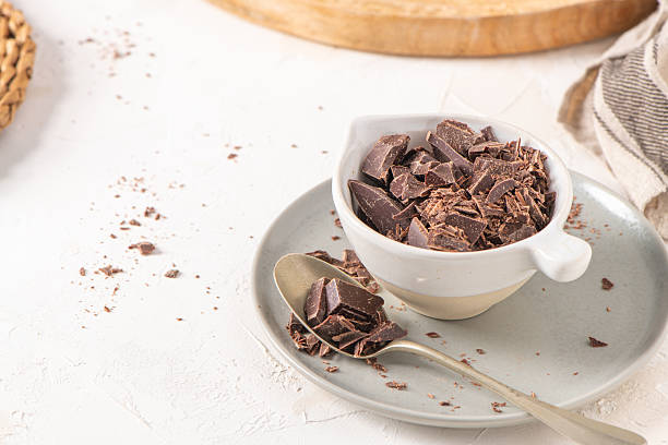 istock Chocolate chunks in a white ceramic cup on a matching saucer with a spoon, set on a textured white surface. The background includes a wooden board 2212125594