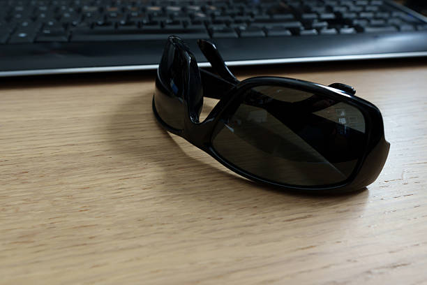 Black sunglasses sit on a wooden desk near a keyboard, suggesting a break from work