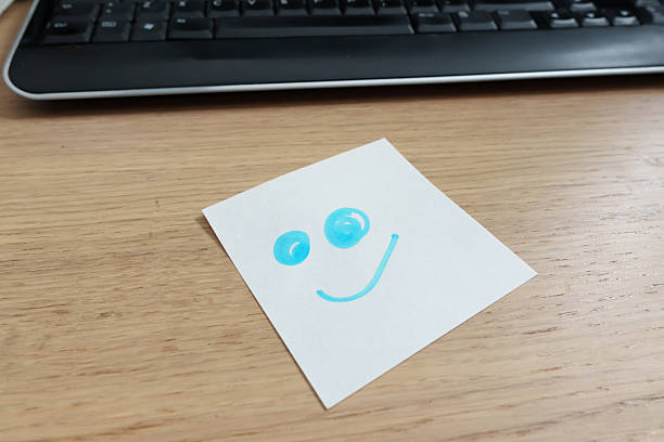 Blue smiley face is drawn on a sticky note placed on a wooden desk with a keyboard in the background