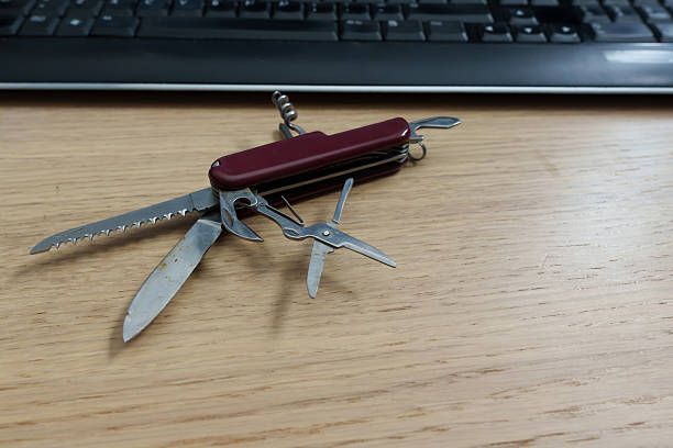 Multifunction red pocket knife unfolded, displaying multiple tools beside computer keyboard on wooden desk surface