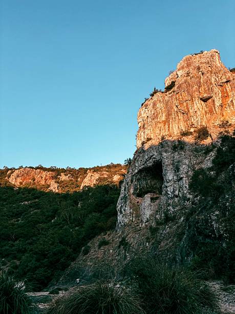 Sunlit rock cliff with a prominent cave opening, surrounded by greenery and contrasting against a vivid blue sky