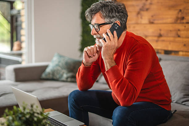 istock Modern senior man sit on sofa with laptop and talk on mobile phone 2212142775