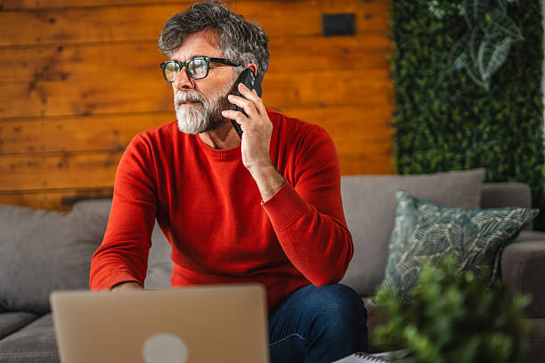 istock Modern senior man sit on sofa with laptop and talk on mobile phone 2212142977