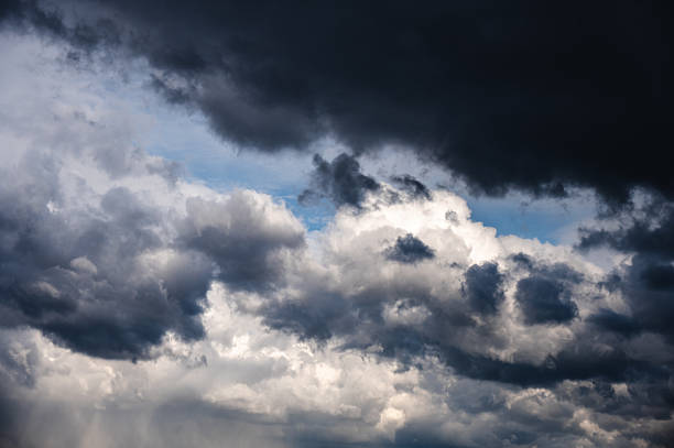 A dramatic scene of dark storm clouds rolling across the sky, partially revealing patches of blue between the heavy, textured layers. The contrast between the light and dark areas evokes a sense of tension and the anticipation of an approaching storm.