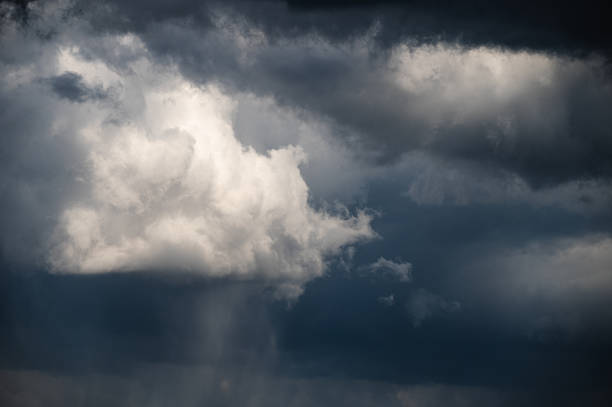 A dramatic scene of dark storm clouds rolling across the sky, partially revealing patches of blue between the heavy, textured layers. The contrast between the light and dark areas evokes a sense of tension and the anticipation of an approaching storm.